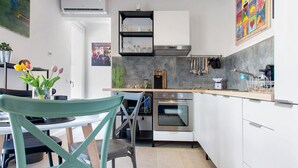Cozinha moderna com armários brancos, forno embutido e exaustor de aço inoxidável. Há uma área de jantar com mesa e cadeiras, e um quadro abstrato colorido na parede.