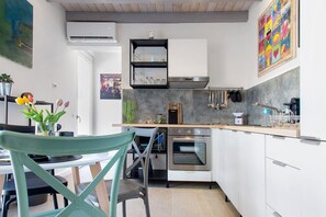 A modern kitchen with white cabinets, a built-in oven, and a stainless steel range hood. There is a dining area with a table and chairs, and a colorful abstract painting on the wall. - 'Residenza La Fonte Dependance' – Perfect for Couples, Private Terrace, Air Conditioning (Laveno-Mombello)
