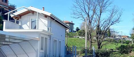 A modern two-story house with a balcony, a paved driveway, and a grassy hill in the background.