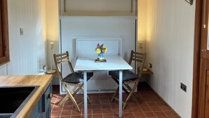 Un pequeño comedor con una mesa blanca, dos sillas de madera, un florero con flores y una lámpara colgante.