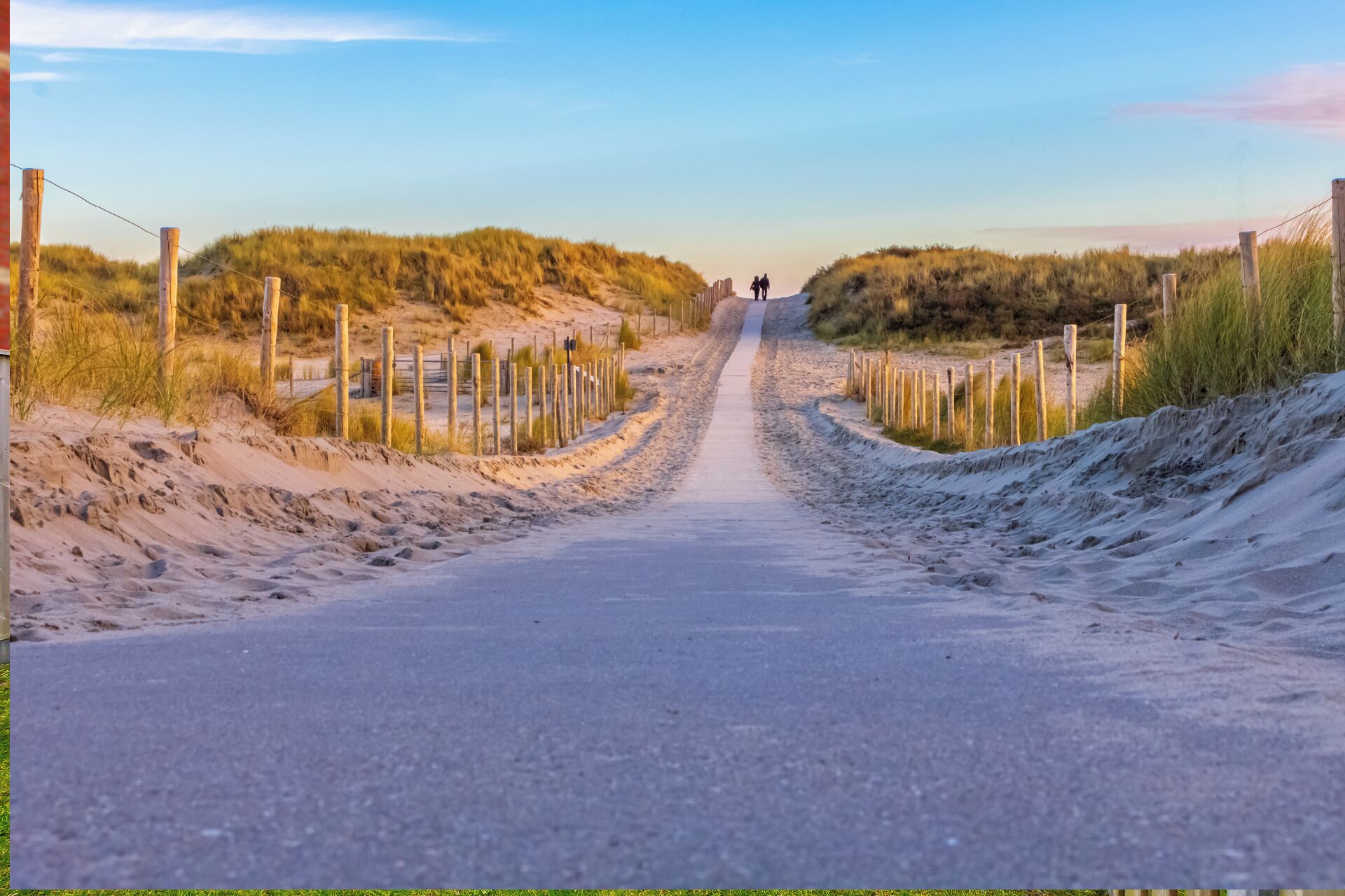 A paved road flanked by wooden fences and sand dunes, leading to a distant point.