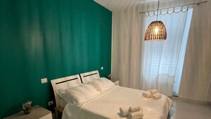 A bedroom with a bed, white bedding, a bedside table, a hanging light fixture, and a window with curtains.