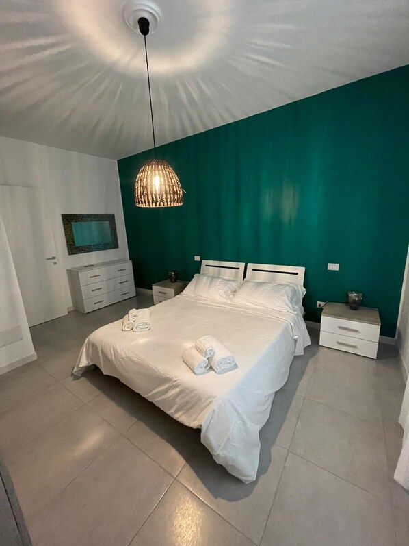 Une chambre moderne avec un grand lit, une literie blanche, un mur d’accent vert, un luminaire suspendu et une petite table de chevet avec une lampe.