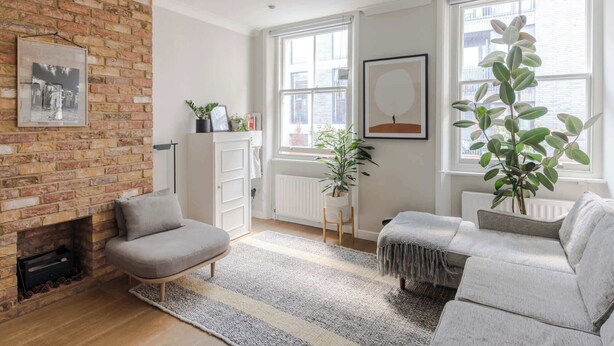 Ein Wohnzimmer mit Backsteinwand, Kamin, einer Couch, einem Sessel und zwei Fenstern mit Pflanzen.