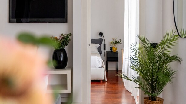 A modern hotel room with a large flat-screen TV, a bed with white bedding, a bedside table with a lamp, and a potted plant. - Garibaldi - Modern 1 BR in Brera by Rentopolis (Milano)