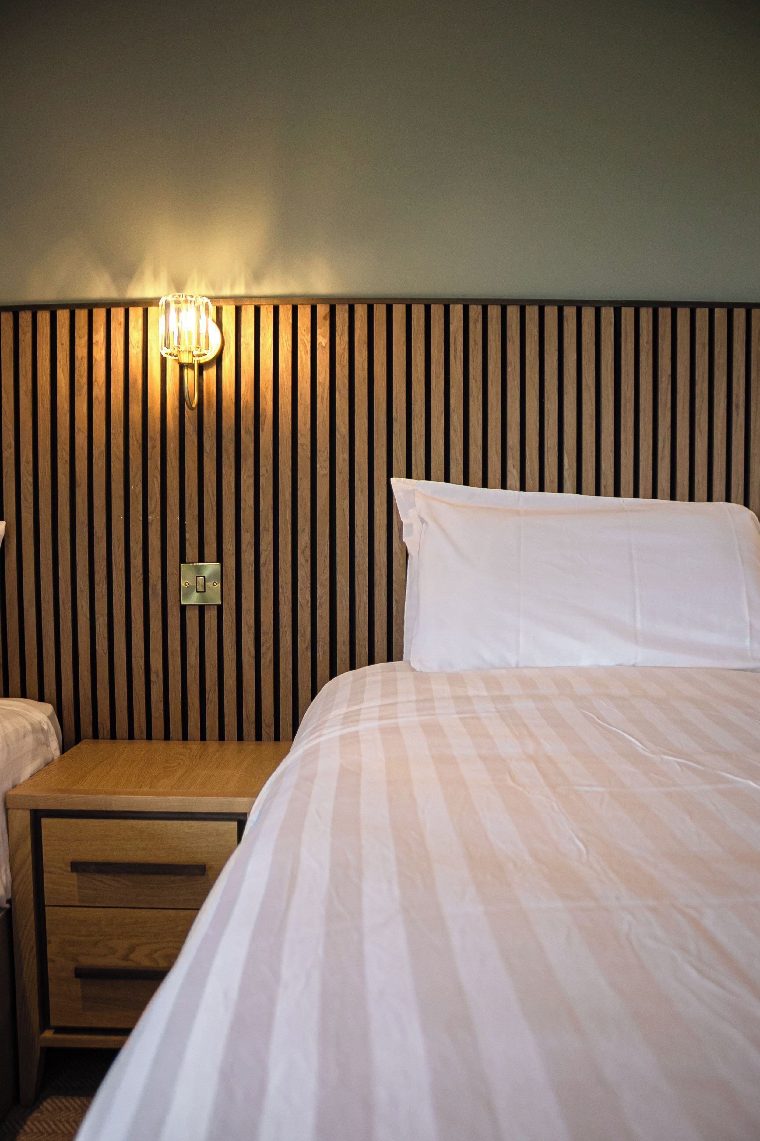A hotel room with a wooden headboard, a bedside table with a drawer, and a bed with white bedding.