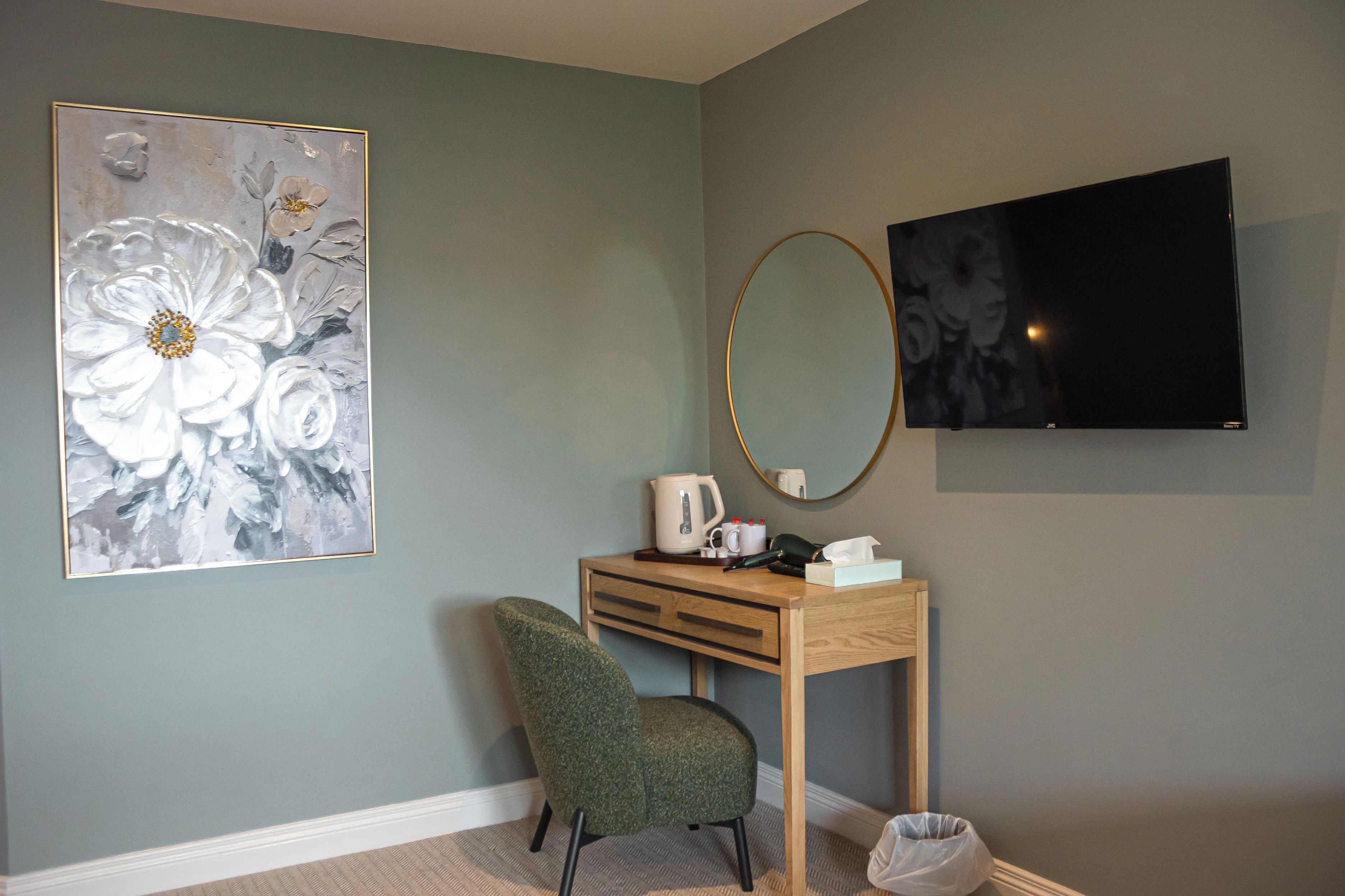 Una habitación con escritorio, silla, televisor de pantalla plana, espejo adosado a la pared y una obra de arte floral de gran tamaño.