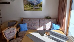 Een woonkamer met een bank, stoelen, een tafel, een klok en een schilderij van een berglandschap.