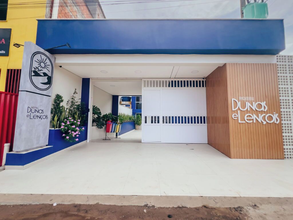 L’entrée d’un hôtel, avec une façade bleue et blanche, un panneau latéral en bois et une enseigne indiquant « Hôtel Dunos e Lencóis ».