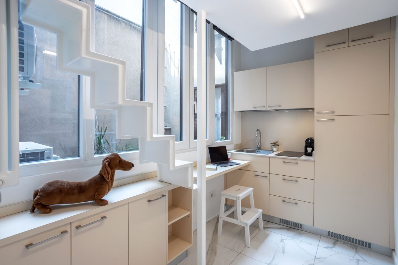Une cuisine moderne avec des meubles blancs, un ordinateur portable posé sur un bureau blanc et un petit tabouret blanc.