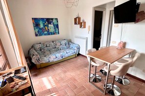 A small living area with a sofa, a table with chairs, a radiator, a TV mounted on the wall, and a shelf with items. - P079 - Porto Recanati, two-room apartment with courtyard in a residence with pool. (Porto Recanati)