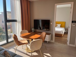 A modern hotel room with a dining area, a bed with a yellow headboard, a TV, and a view of the city. - The Sky (Phu Quoc)