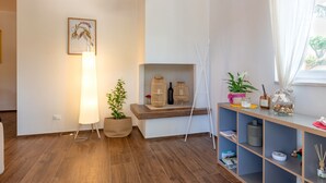 Una habitación moderna con piso de madera, una lámpara de pie, una planta en maceta, un estante con objetos decorativos y una ventana con cortinas.