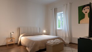 Un dormitorio con cama, mesita de noche, lámpara, ventana con cortinas y un cuadro en la pared.