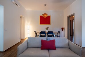 Una sala de estar con un sofá, una mesa de comedor con sillas, una obra de arte con marco rojo y un jarrón con flores.