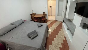 Ett hotellrum med en säng, ett litet matbord med stolar, en tv som är monterad på ett vitt skåp och en dörr som leder ut till en hall.