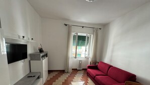 Ett vardagsrum med en röd soffa, en tv som är monterad på väggen, ett litet bord och ett fönster med gardiner.