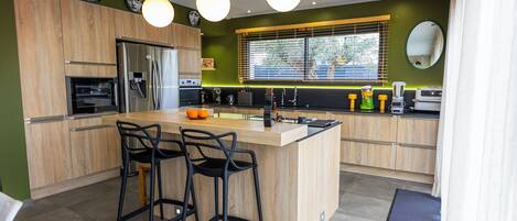 Une cuisine moderne avec des meubles en bois, un îlot central doté de tabourets de bar, un four encastré et une grande fenêtre avec des stores.