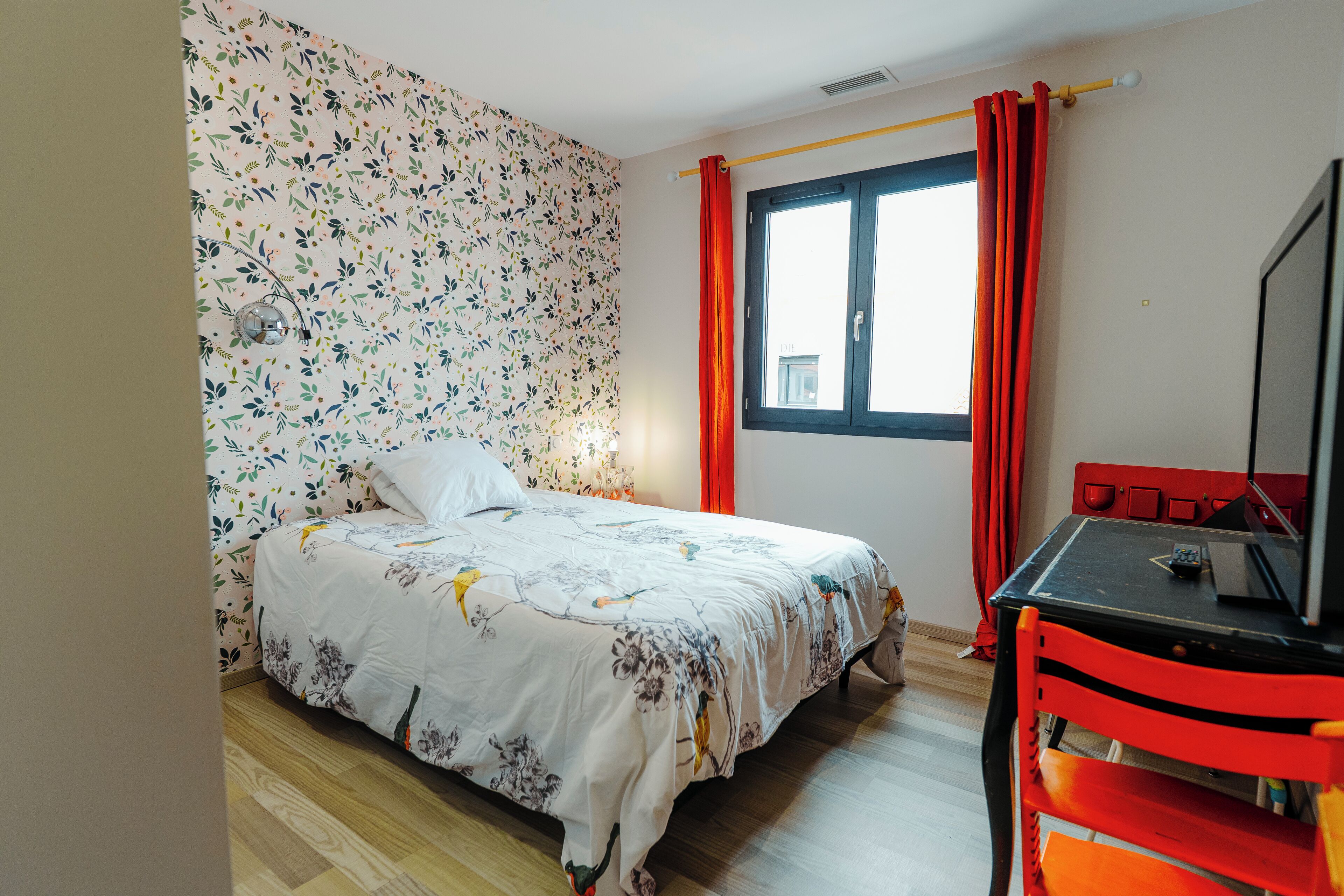 A bedroom with a bed, a desk, a chair, a window with curtains, and a wall with a floral pattern.