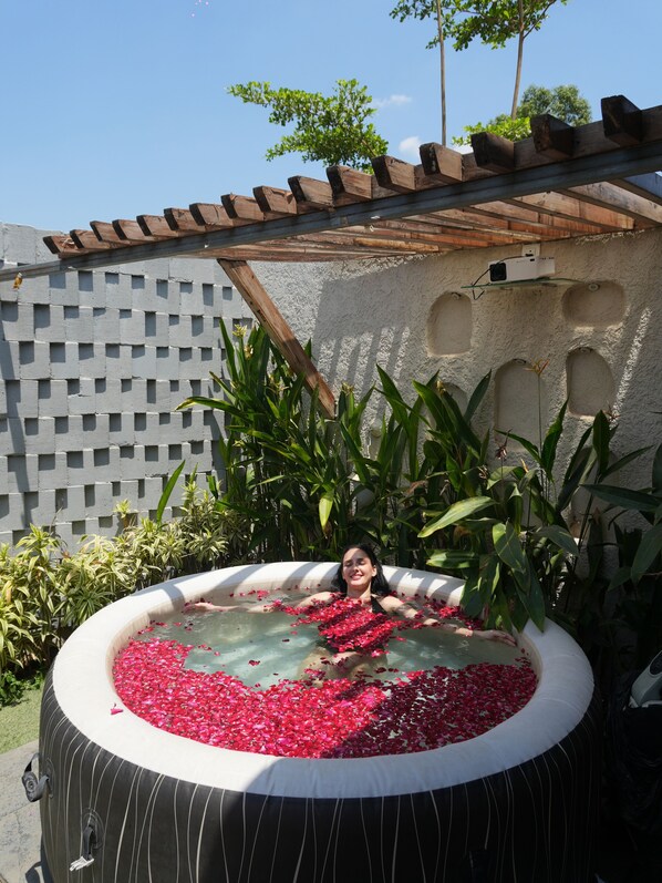 A person in a hot tub filled with rose petals, surrounded by greenery and a pergola.