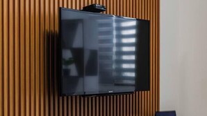 Un salón moderno con un televisor de pantalla plana montado en una pared de madera, un mueble de entretenimiento gris con un control remoto, y un objeto azul pequeño sobre el mueble.