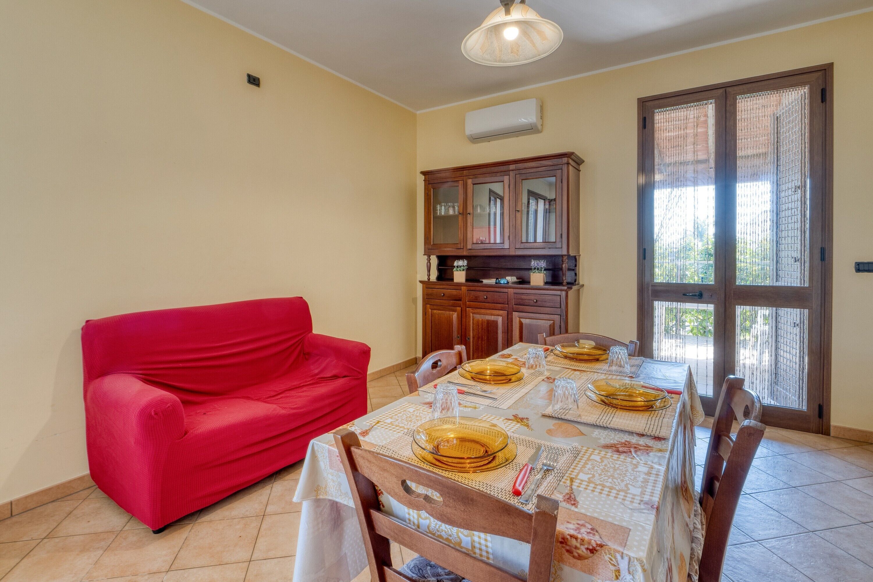 Un coin repas avec une table dressée pour quatre personnes, un canapé rouge, des meubles en bois et une porte-fenêtre coulissante.