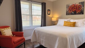A bedroom with a large bed, a red armchair, a window with blinds, a painting on the wall, and a bedside lamp. - Lazy Hiker by Chatuge Home Concierge | Walk to Downtown Hayesville | Fire Pit | Pet Friendly (Hayesville)
