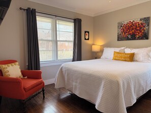 A bedroom with a large bed, a red armchair, a window with blinds, a painting on the wall, and a bedside lamp.
