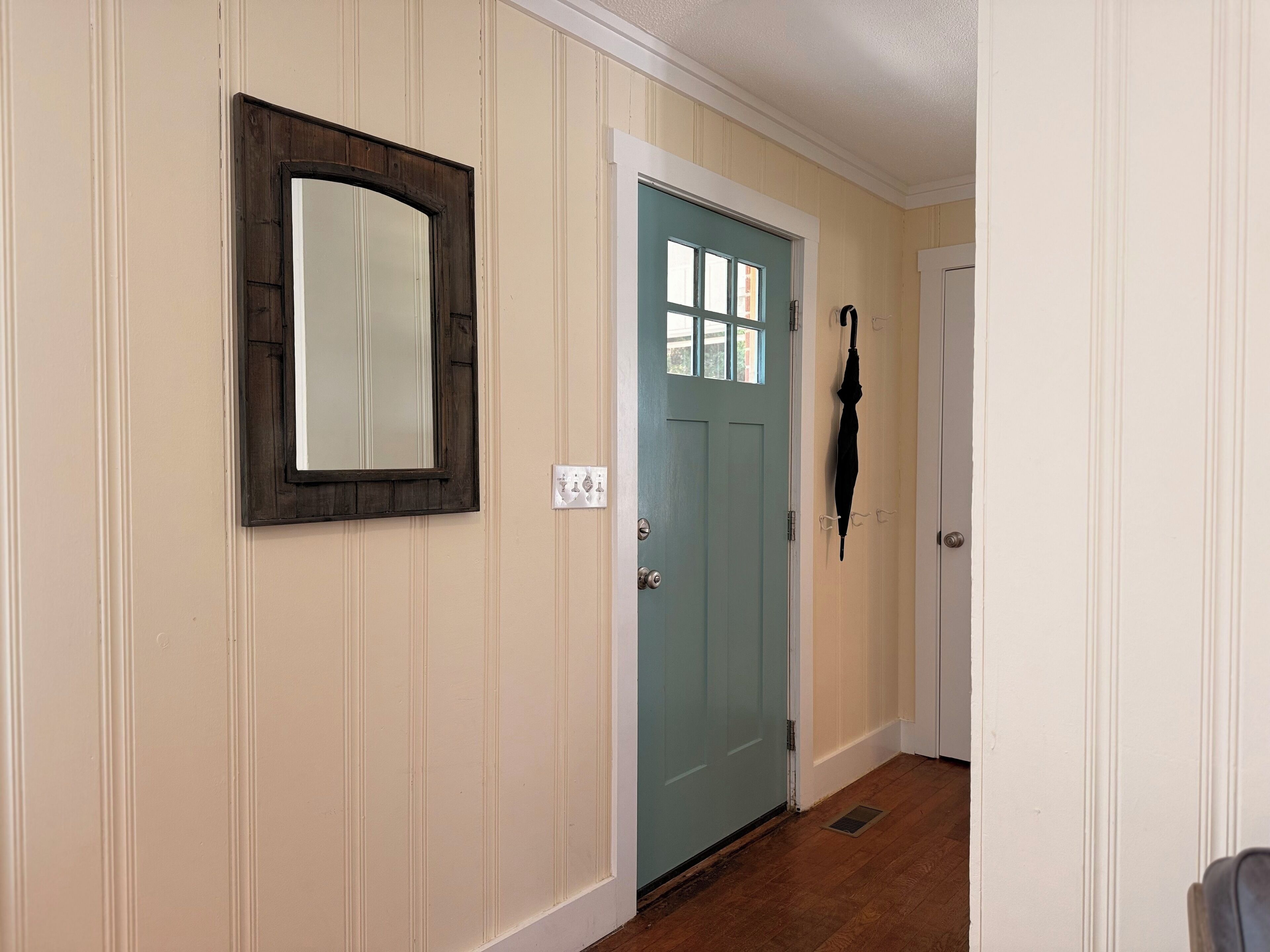 A hallway with a light blue door, a dark wooden-framed mirror, and a black umbrella hanging on the wall.