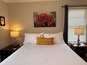 A neatly made bed with white bedding, a single decorative pillow with the text "Together our favorite place," a painting of red flowers on the wall, and two bedside lamps.