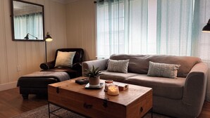 A living room with a sofa, a chair, a wooden coffee table, and a mirror on the wall. - Lazy Hiker by Chatuge Home Concierge | Walk to Downtown Hayesville | Fire Pit | Pet Friendly (Hayesville)
