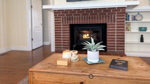 A cozy living room with a brick fireplace, a wooden coffee table with candles, a potted plant, and a book titled "The Whisky Book". - Lazy Hiker by Chatuge Home Concierge | Walk to Downtown Hayesville | Fire Pit | Pet Friendly (Hayesville)