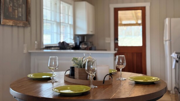 A round wooden dining table with green plates and wine glasses, set in a room with a kitchen in the background. - Lazy Hiker by Chatuge Home Concierge | Walk to Downtown Hayesville | Fire Pit | Pet Friendly (Hayesville)