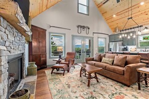 Un salon chaleureux avec une cheminée en pierre, un plafond en bois et une vue sur l’extérieur grâce à de grandes fenêtres.