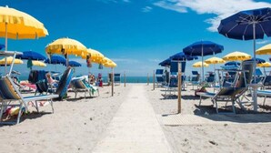 Ein Strand mit Reihen von Liegestühlen und Sonnenschirmen, einer Holzpromenade und dem Meer im Hintergrund.
