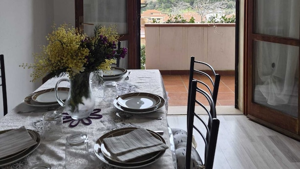 A dining area with a table set for four, a vase with flowers, and a view of a mountainous landscape through open doors. - Three-room apartment Nicole with private garden Elba Island (Porto Azzurro)