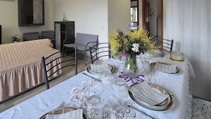 A dining area with a table set for four, a bed with a beige cover, a television, and a door leading to another room. - Three-room apartment Nicole with private garden Elba Island (Porto Azzurro)