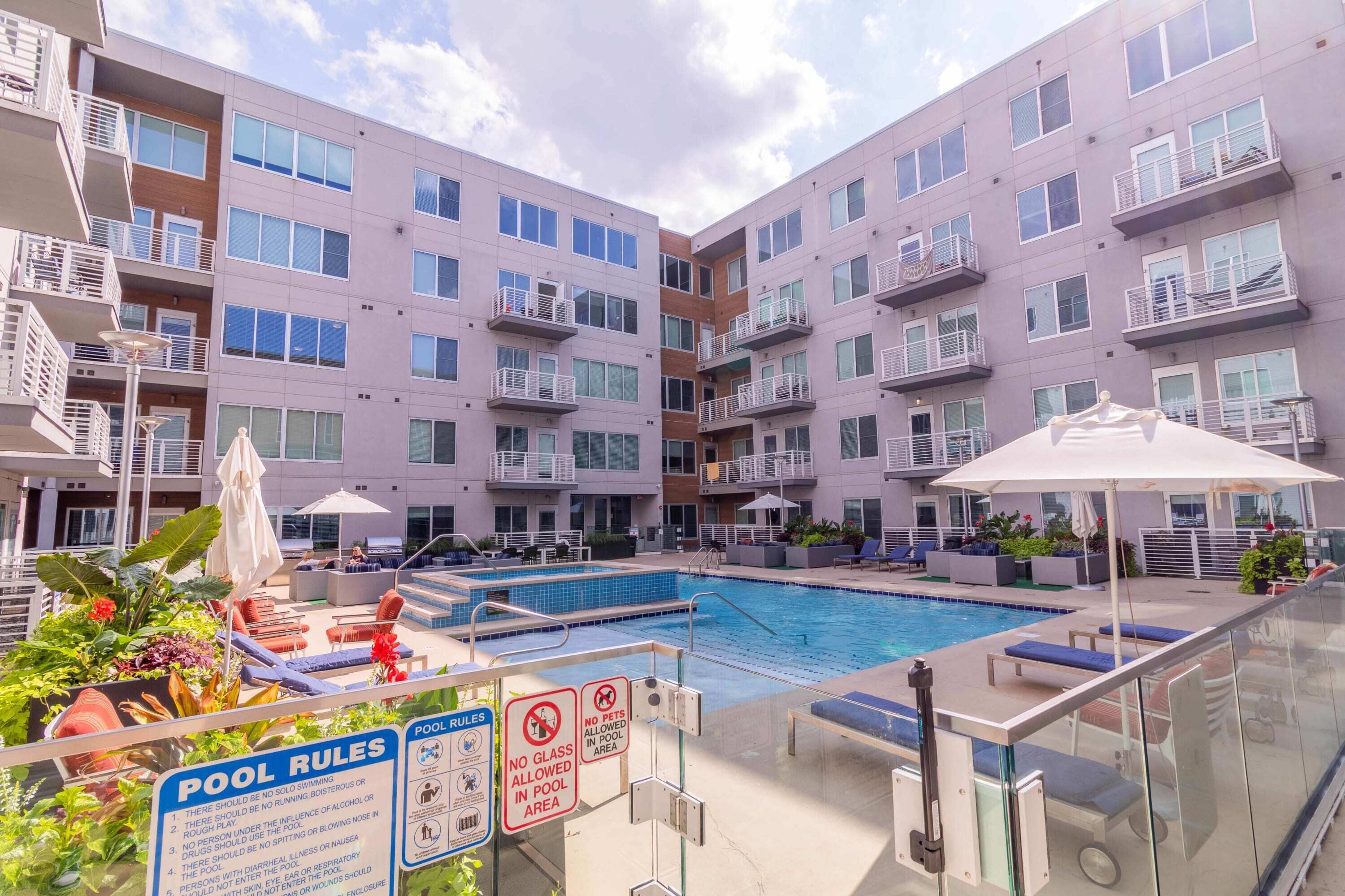 A pool area with clear pool rules signs, lounge chairs, and a modern apartment building in the background.