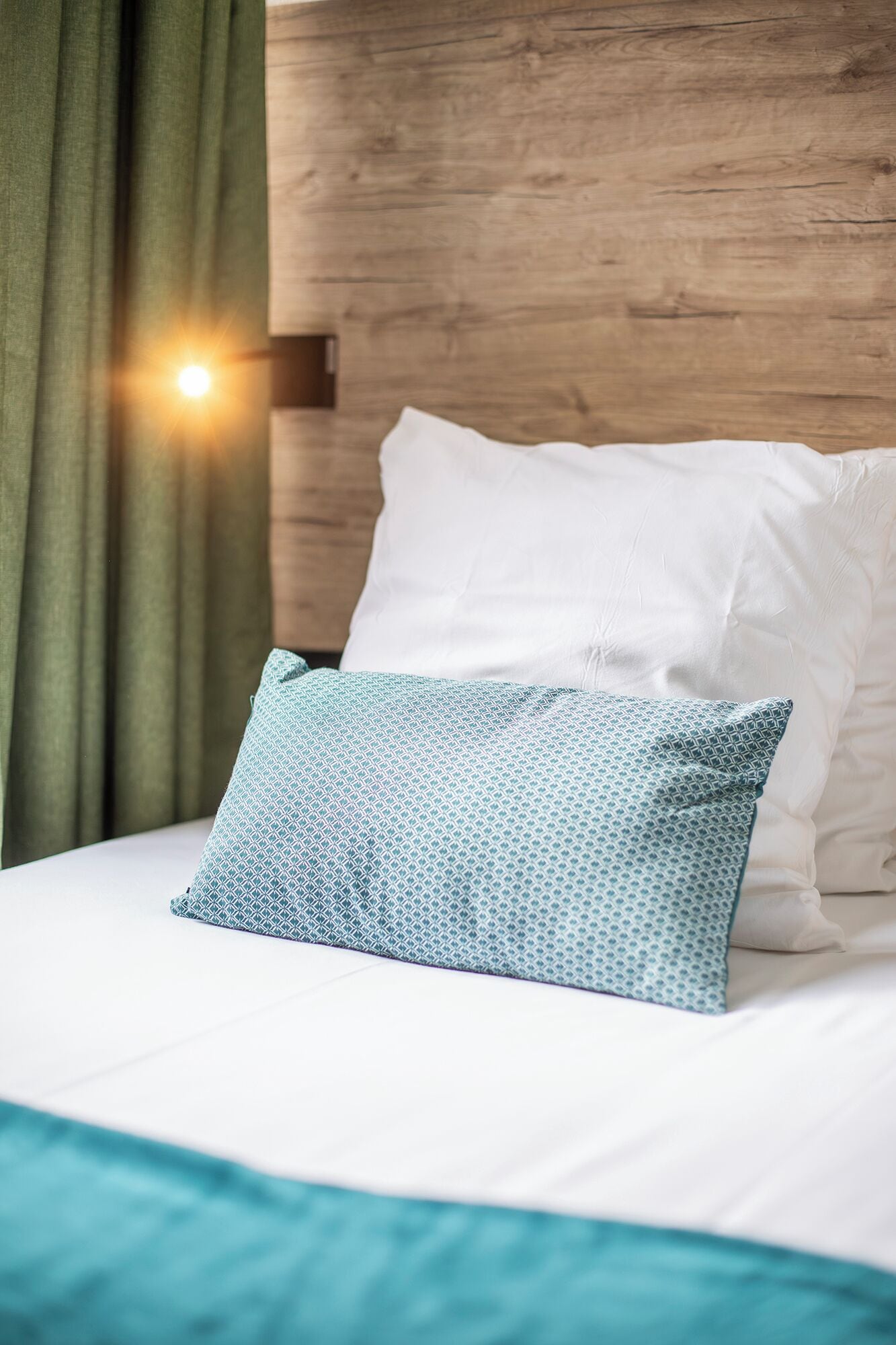 A bed with white bedding and a patterned pillow, a wooden headboard, and a green curtain.