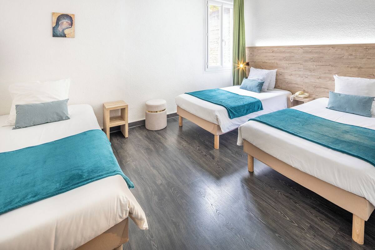 Une chambre d’hôtel avec trois lits, un petit tabouret en bois, un pouf rond et un tableau accroché au mur.