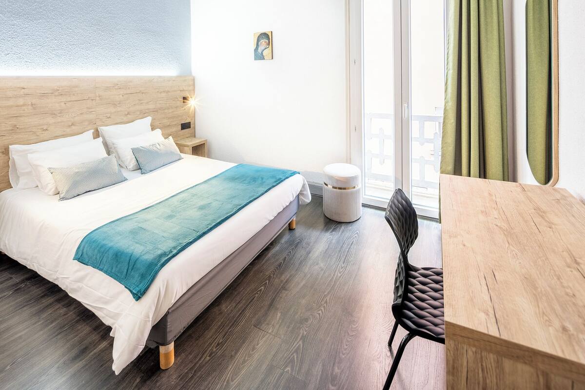 Une chambre d’hôtel avec un lit, une petite table, une chaise, un pouf rond et une fenêtre avec des rideaux.