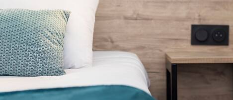 A neatly made bed with a teal bedspread and patterned pillows, a wooden headboard, and a bedside table with a speaker and a light.