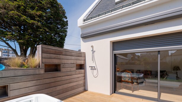 A modern outdoor hot tub area with a wooden deck, a glass door leading to a living room, and a large tree in the background. - Maison Desmathi: terrace with jacuzzi, garden, in Saint-Pierre-Quiberon (Saint-Pierre-Quiberon)