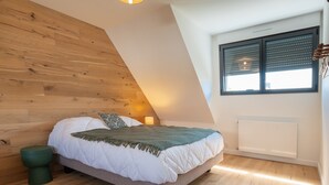 A bedroom with a sloped ceiling, a bed with white bedding, a green throw blanket, a bedside table with a lamp, and a window with blinds. - Maison Desmathi: terrace with jacuzzi, garden, in Saint-Pierre-Quiberon (Saint-Pierre-Quiberon)
