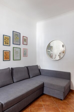 Ein graues Ecksofa an einer weißen Wand, mit sechs gerahmten Kunstwerken und einem runden Spiegel.