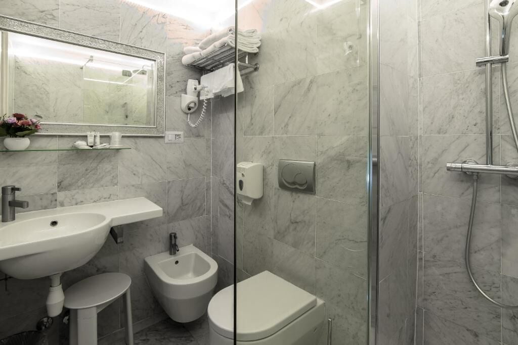 A modern bathroom with a marble sink, bidet, and toilet, a glass shower enclosure, and a towel rack with towels.