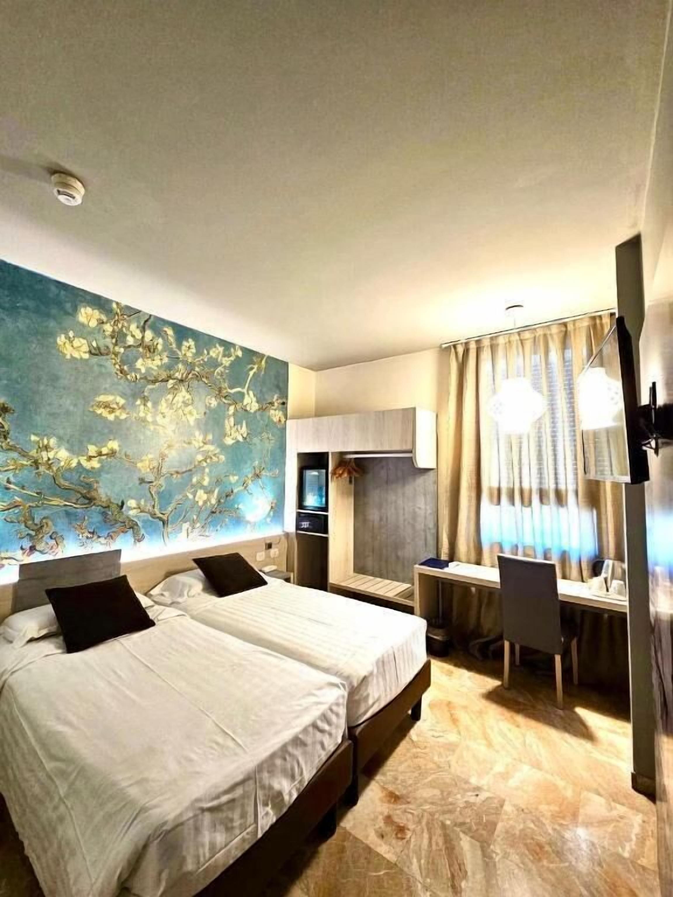 A hotel room with two beds, a desk, a TV, and a large floral mural on the wall.