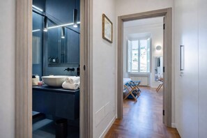 A modern bathroom with a large mirror, a sink, and a towel rack. A hallway leads to a bedroom with a bed and a window. - LVD Lucina Luxury Apartament al Centro di Roma (Rome)