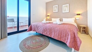 Un dormitori amb un llit, un petit tamboret, una làmpada penjada a la paret i una vista a través d'una porta de vidre.