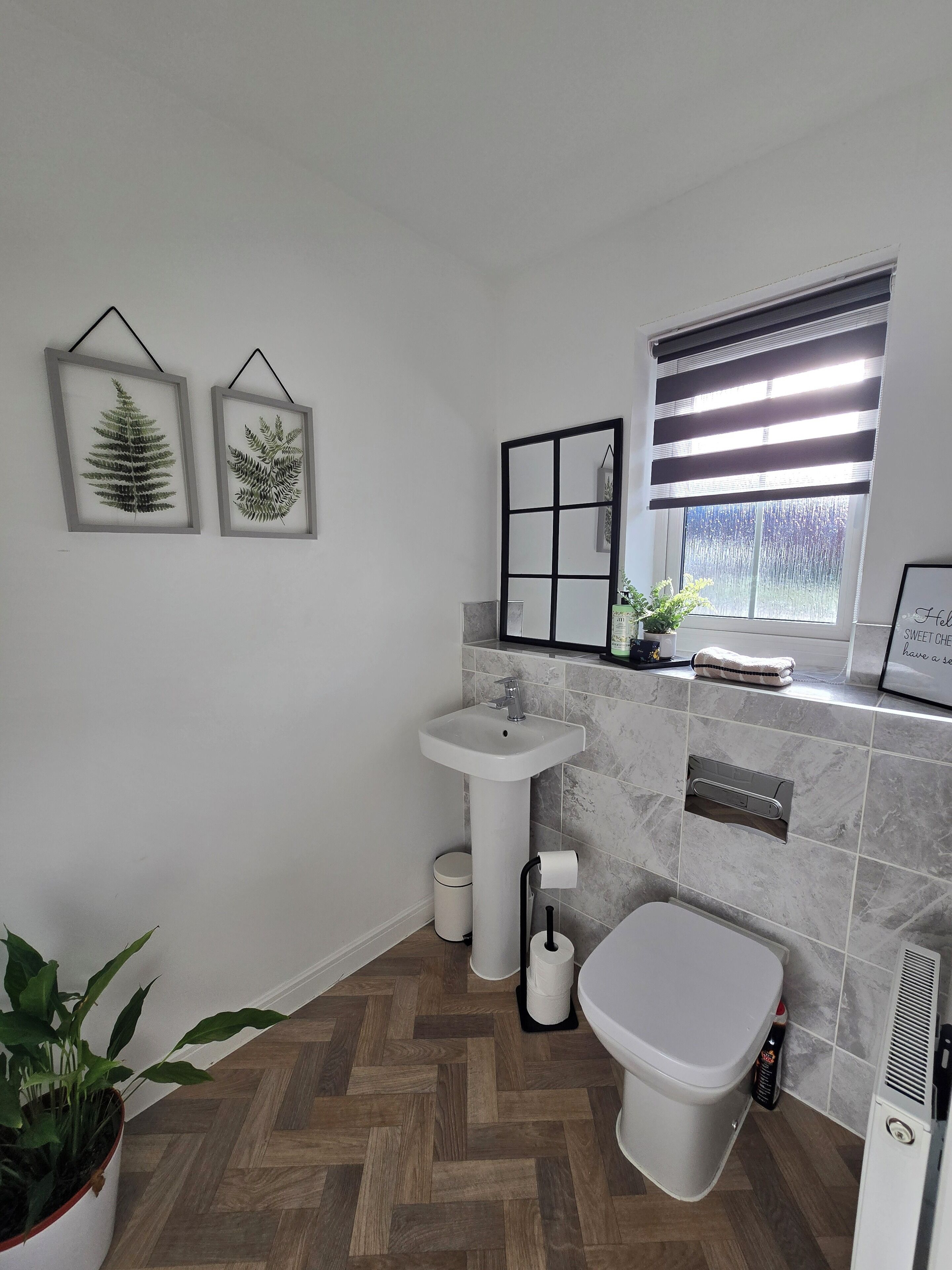 Ein modernes Badezimmer mit einem weißen Waschbecken, einer Toilette und einem Fischgrätparkettboden. An den Wänden befinden sich botanische Drucke in Rahmen, und es gibt ein Fenster mit einem schwarzen Rollo.